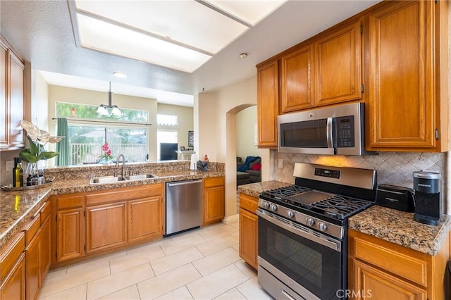 kitchen featuring arched walkways, backsplash, appliances with stainless steel finishes, brown cabinetry, and a sink