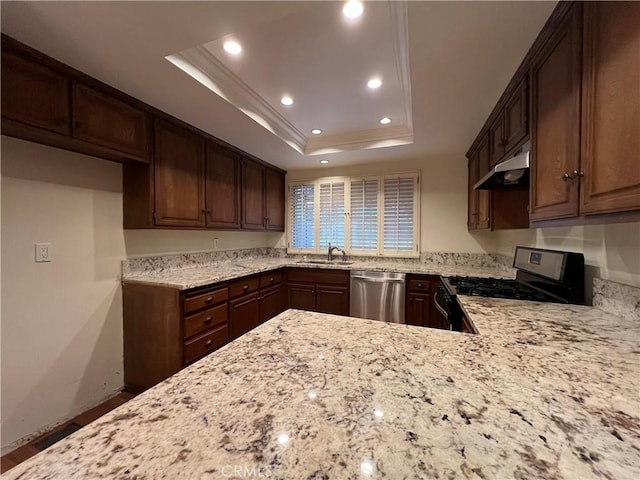 kitchen with ornamental molding, under cabinet range hood, a tray ceiling, light stone counters, and appliances with stainless steel finishes