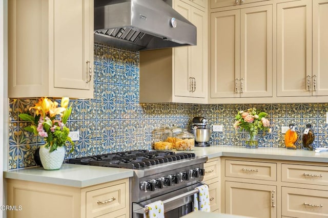 kitchen with decorative backsplash, stainless steel range, cream cabinetry, and wall chimney exhaust hood