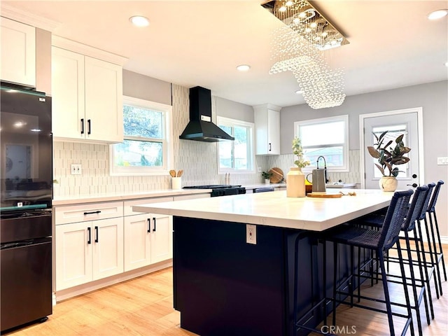 kitchen featuring wall chimney range hood, a center island, white cabinets, decorative light fixtures, and light wood-type flooring