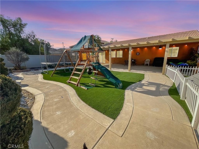 playground at dusk featuring a trampoline, a yard, and a patio