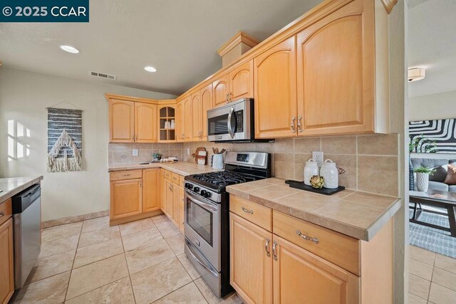 kitchen with light tile patterned flooring, appliances with stainless steel finishes, tile countertops, light brown cabinetry, and tasteful backsplash