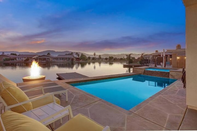 pool at dusk featuring an in ground hot tub, a water view, a patio, and a fire pit