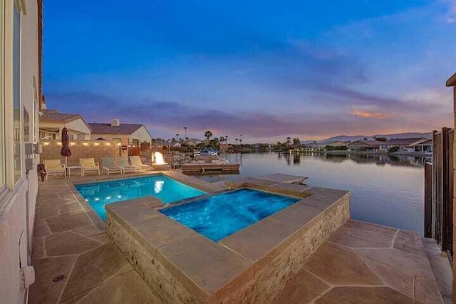 pool at dusk with an in ground hot tub, a water view, and a patio