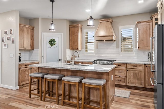 kitchen with pendant lighting, premium range hood, stainless steel appliances, light stone counters, and a center island with sink