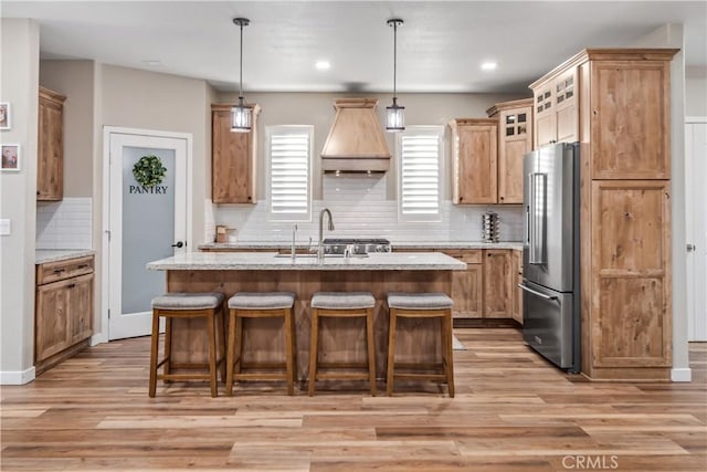 kitchen with high end fridge, sink, hanging light fixtures, an island with sink, and custom range hood