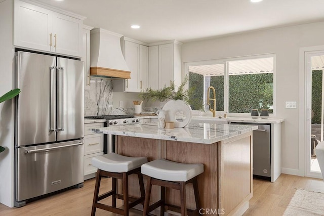 kitchen with custom exhaust hood, white cabinetry, light stone counters, appliances with stainless steel finishes, and a kitchen island