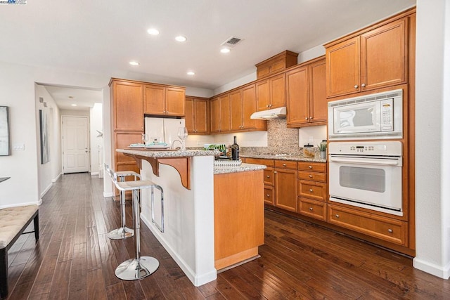 kitchen with a kitchen breakfast bar, light stone counters, dark wood-type flooring, a center island with sink, and white appliances