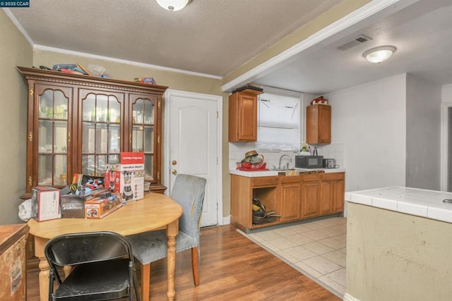 kitchen with decorative backsplash, ornamental molding, tile counters, light hardwood / wood-style floors, and a textured ceiling