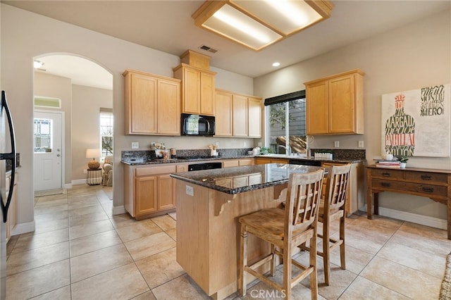 kitchen with dark stone countertops, a center island, black appliances, light tile patterned flooring, and light brown cabinets