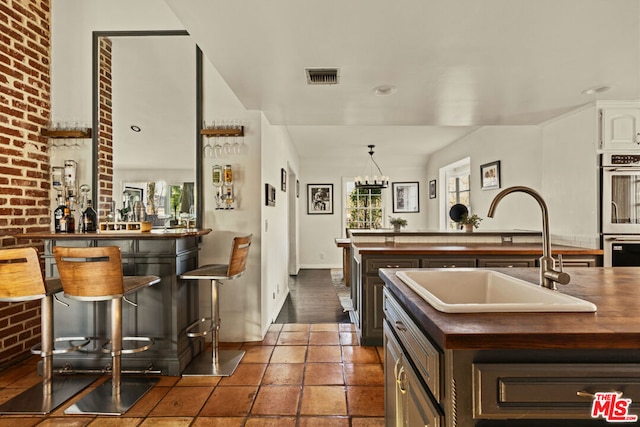 kitchen with brick wall, sink, stainless steel double oven, dark brown cabinetry, and a center island with sink