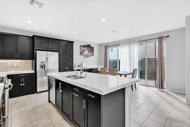 kitchen with sink, a center island with sink, light tile patterned floors, stainless steel appliances, and decorative backsplash