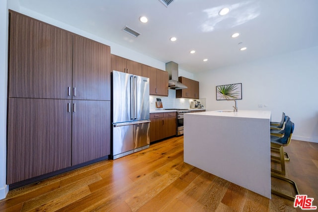 kitchen with an island with sink, a breakfast bar area, hardwood / wood-style flooring, high end appliances, and wall chimney exhaust hood