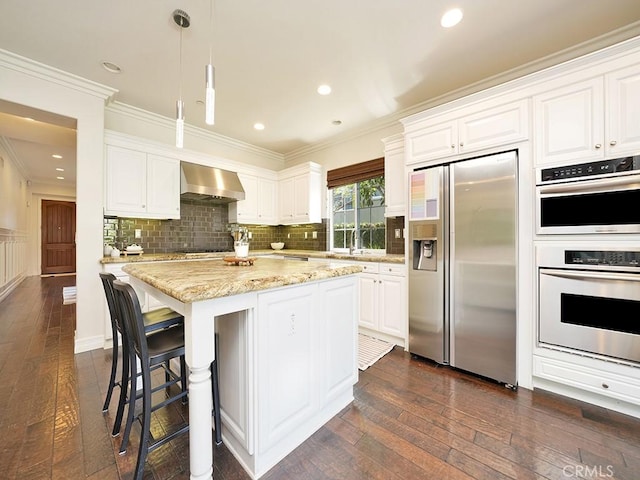 kitchen featuring white cabinetry, stainless steel fridge, hanging light fixtures, light stone counters, and wall chimney range hood