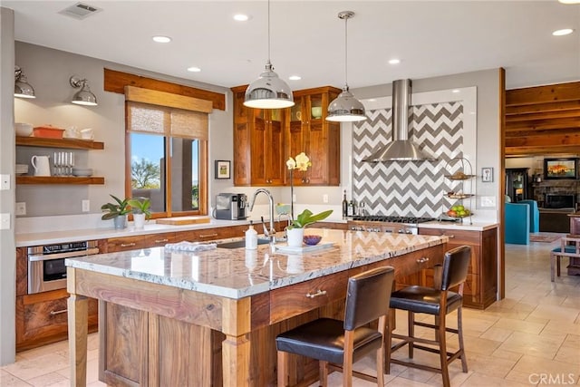 kitchen with light stone counters, hanging light fixtures, an island with sink, stainless steel appliances, and wall chimney range hood