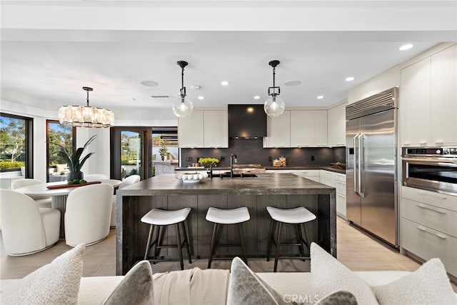 kitchen with appliances with stainless steel finishes, sink, wall chimney exhaust hood, hanging light fixtures, and an island with sink