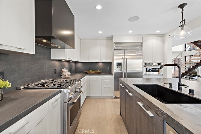 kitchen with wall chimney range hood, premium appliances, white cabinetry, and sink