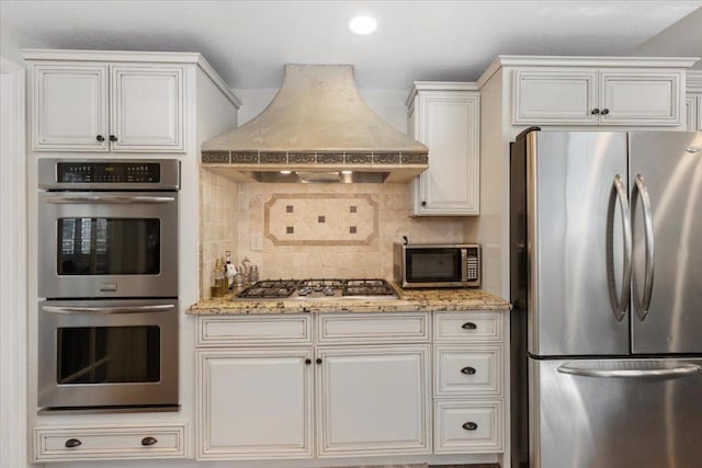 kitchen with custom exhaust hood, white cabinetry, light stone counters, appliances with stainless steel finishes, and backsplash