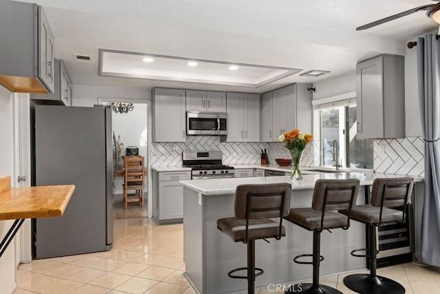 kitchen featuring a breakfast bar area, gray cabinetry, appliances with stainless steel finishes, a tray ceiling, and kitchen peninsula