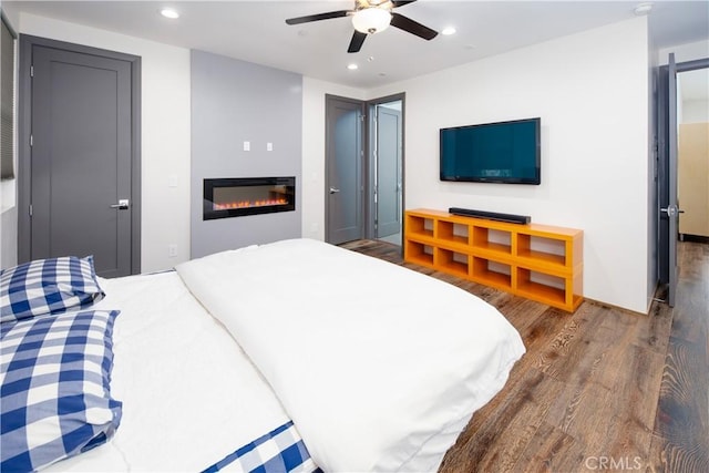 bedroom with a ceiling fan, a glass covered fireplace, dark wood-type flooring, and recessed lighting