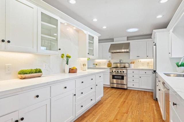 kitchen with wall chimney range hood, sink, light hardwood / wood-style flooring, white cabinetry, and stainless steel appliances