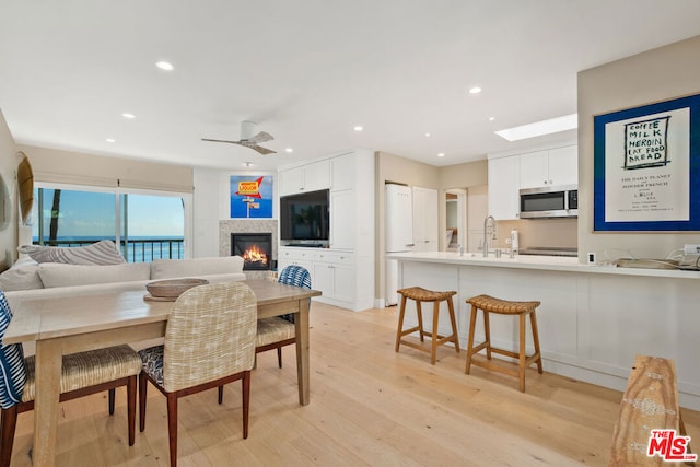 dining room with sink, a skylight, ceiling fan, a tiled fireplace, and light hardwood / wood-style floors