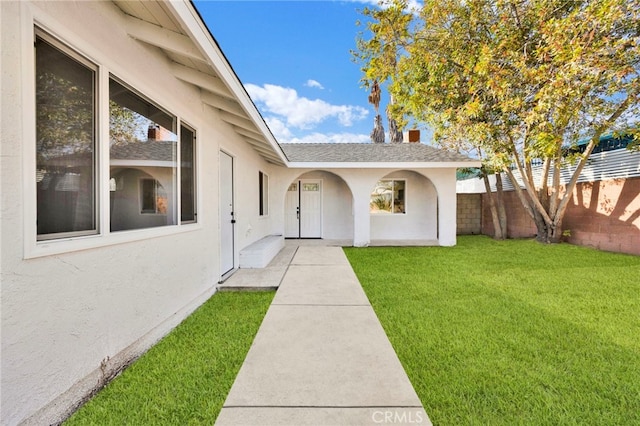 property entrance featuring a shingled roof, fence, a lawn, and stucco siding