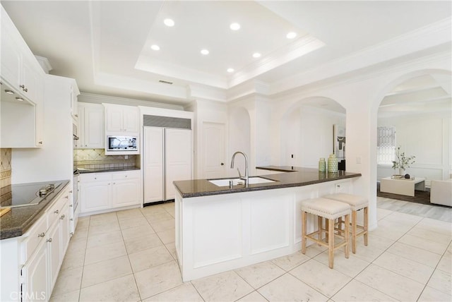 kitchen featuring backsplash, built in appliances, white cabinets, a center island with sink, and a raised ceiling