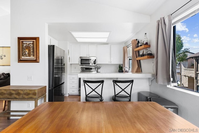 kitchen with stainless steel appliances, white cabinetry, lofted ceiling with skylight, and tile countertops