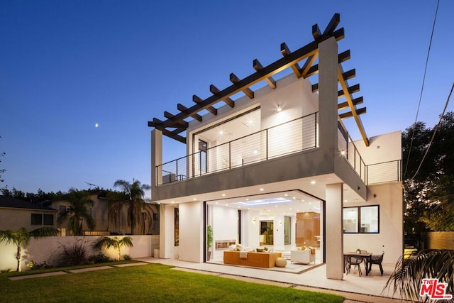 back house at dusk with a balcony, outdoor lounge area, a patio, and a lawn