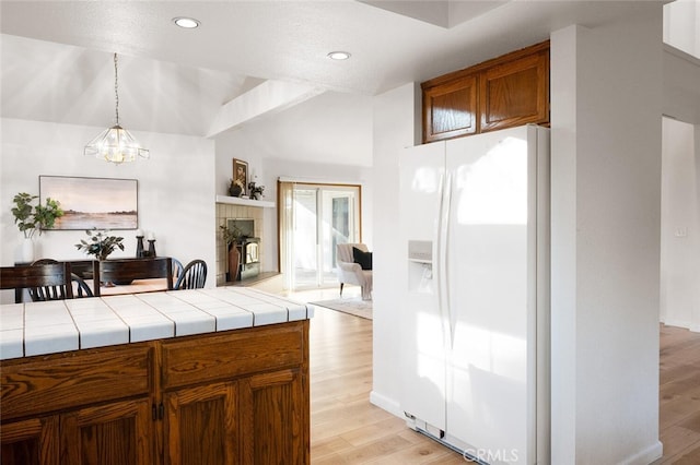 kitchen featuring brown cabinets, tile counters, light wood-style flooring, a tiled fireplace, and white fridge with ice dispenser