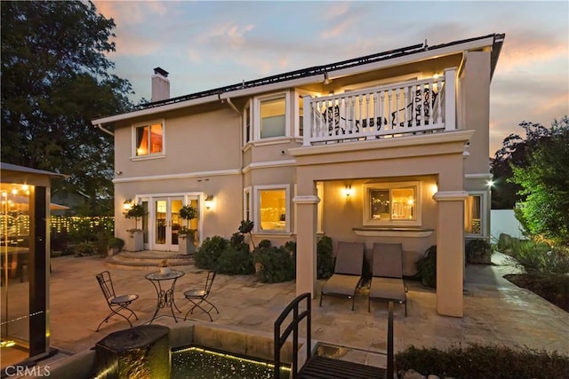 back of house at dusk featuring stucco siding, french doors, a chimney, a balcony, and a patio area