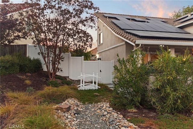 property exterior at dusk with a tile roof, fence, roof mounted solar panels, and stucco siding