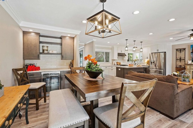 dining area featuring ornamental molding, beverage cooler, ceiling fan with notable chandelier, and light wood-type flooring