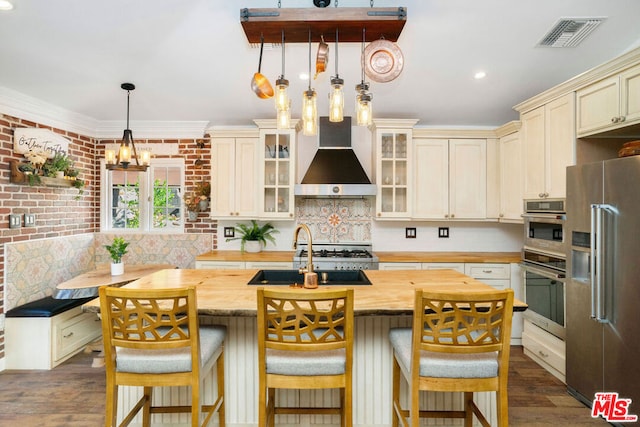 kitchen featuring sink, a center island with sink, appliances with stainless steel finishes, breakfast area, and wall chimney range hood