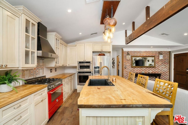 kitchen featuring appliances with stainless steel finishes, an island with sink, butcher block counters, sink, and wall chimney exhaust hood