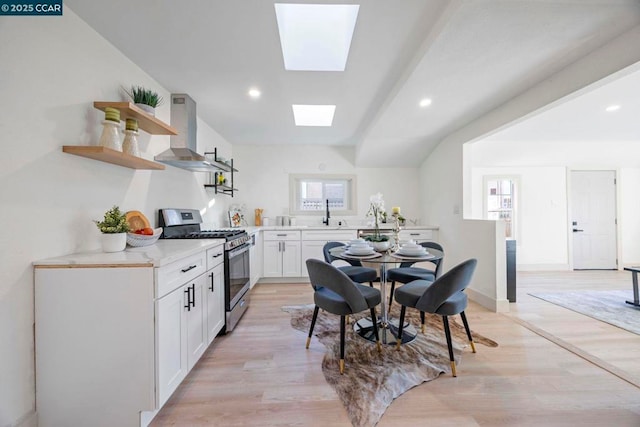 kitchen featuring wall chimney exhaust hood, stainless steel gas range, white cabinetry, a skylight, and light hardwood / wood-style floors