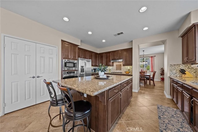 kitchen with appliances with stainless steel finishes, backsplash, dark brown cabinets, a center island, and light stone countertops