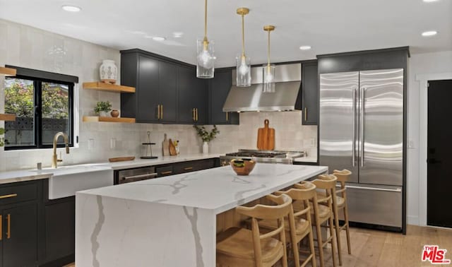 kitchen featuring pendant lighting, appliances with stainless steel finishes, a center island, light stone counters, and wall chimney exhaust hood