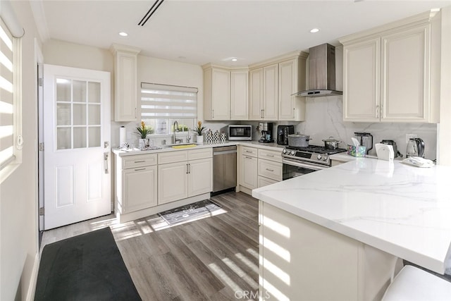 kitchen with sink, appliances with stainless steel finishes, kitchen peninsula, light stone countertops, and wall chimney range hood