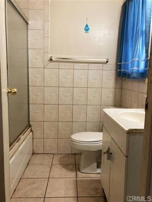 full bathroom with bath / shower combo with glass door, tile walls, tile patterned flooring, vanity, and toilet