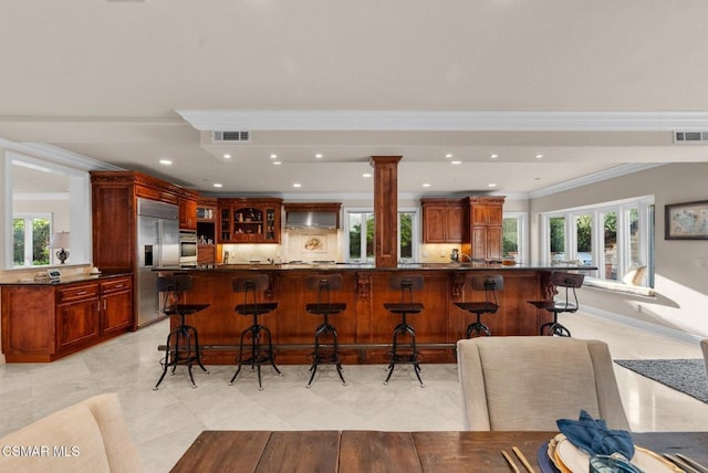 kitchen with crown molding, a breakfast bar, appliances with stainless steel finishes, wall chimney exhaust hood, and ornate columns