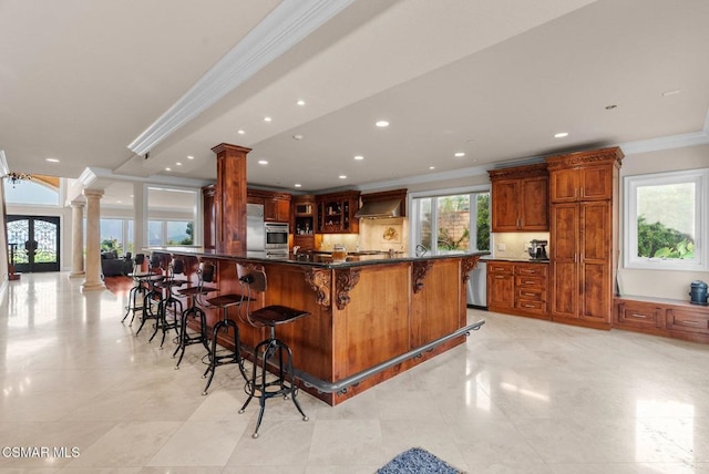 kitchen with crown molding, appliances with stainless steel finishes, a kitchen breakfast bar, decorative columns, and wall chimney range hood