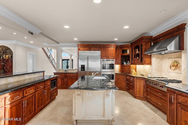 kitchen featuring built in appliances, a center island with sink, ornamental molding, dark stone counters, and wall chimney range hood