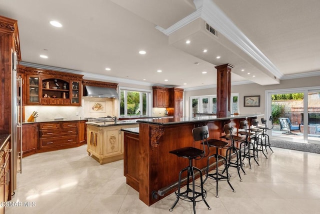 kitchen featuring a breakfast bar area, ornate columns, a center island with sink, a healthy amount of sunlight, and wall chimney range hood