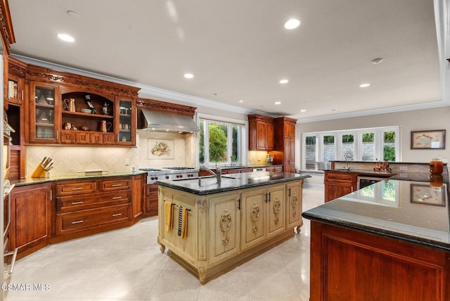 kitchen featuring wall chimney range hood, decorative backsplash, dark stone counters, and a center island with sink