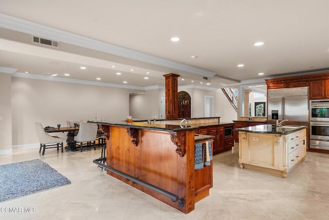 kitchen featuring crown molding, a breakfast bar, a kitchen island with sink, decorative columns, and built in appliances