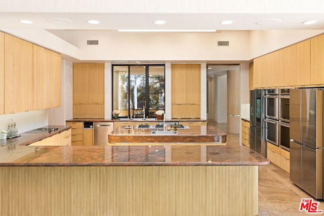 kitchen with light brown cabinets, kitchen peninsula, dark stone counters, and appliances with stainless steel finishes