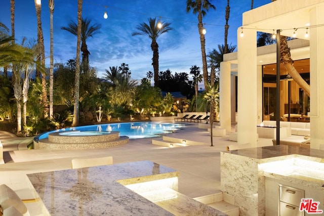 pool at dusk featuring a community hot tub, a patio, and exterior kitchen