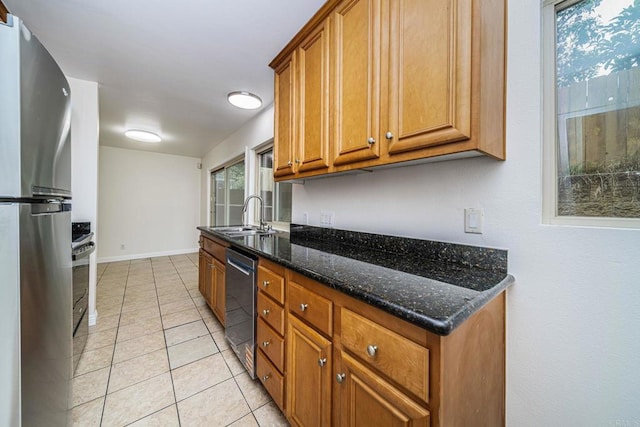kitchen with a sink, appliances with stainless steel finishes, brown cabinets, light tile patterned floors, and dark stone counters